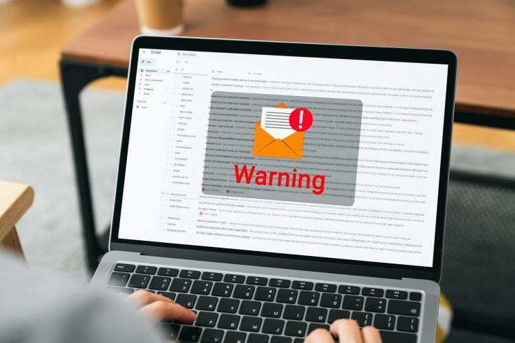 Person tippt auf Laptop, auf dem Bildschirm ist eine E-Mail-Oberfläche mit einem großen Warnsymbol über einem Umschlag zu sehen – Hinweis auf eine mögliche Phishing-Mail.