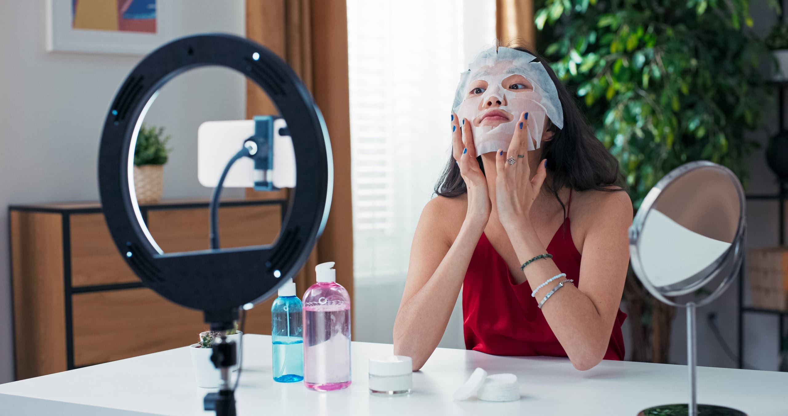 Influencerin demonstriert eine Gesichtspflege-Maske vor der Kamera und nimmt mit Ringlicht und Smartphone ein Video für Social Media auf.
