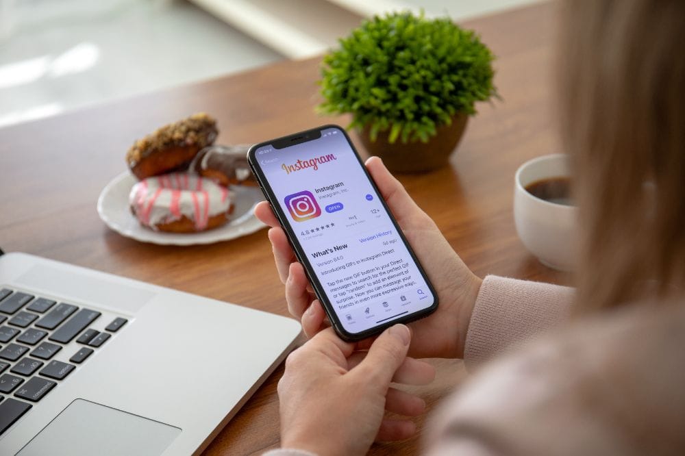 Person hält Smartphone mit geöffneter Instagram-App-Seite im App Store. Auf dem Tisch stehen ein Laptop, eine Tasse Kaffee und ein Teller mit Donuts.