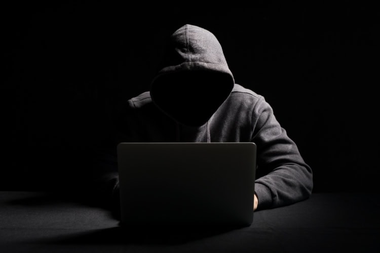 Dunkel gekleidete, gesichtslose Person mit Kapuze sitzt vor einem Laptop in einem dunklen Raum – symbolische Darstellung eines Hackers.