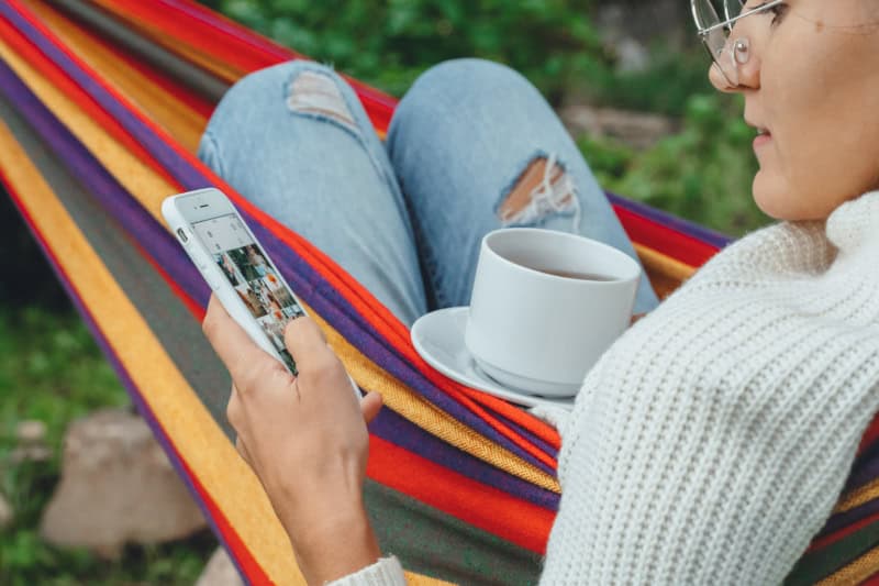 Frau liegt in einer bunten Hängematte, hält ein Smartphone in der einen Hand und eine Tasse Tee in der anderen.