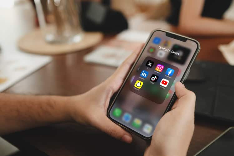 Ein Mann hält ein Smartphone. Das Display zeigt die Social Media Apps X, Instagram, Facebook, LinkedIn, TikTok, YouTube und Snapchat.